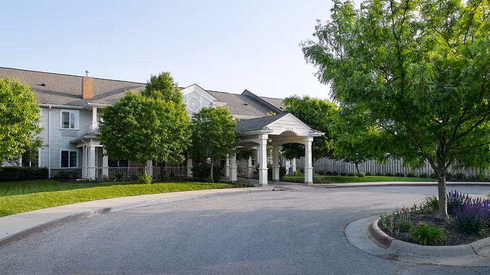 Exterior view of a senior living facility named Celebration Villa of Hearthstone, showing a large building with a covered entrance, surrounded by green trees and landscaping under a clear sky.