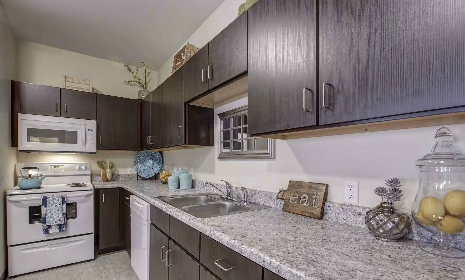 Modern kitchen with dark wood cabinets, a white stove with a blue kettle, a white microwave, a double sink, and a granite countertop. Decorative items include a blue plate, jars, a small wooden sign that says 'eat', a silver vase with a plant, and a glass jar with lemons.