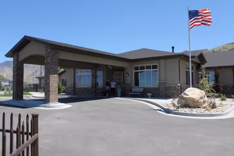 Exterior view of a single-story assisted living facility building with a covered entrance supported by stone pillars. There is an American flag on a flagpole near the entrance, a large rock with landscaping, and a few people standing under the covered area. Mountains are visible in the background under a clear blue sky.