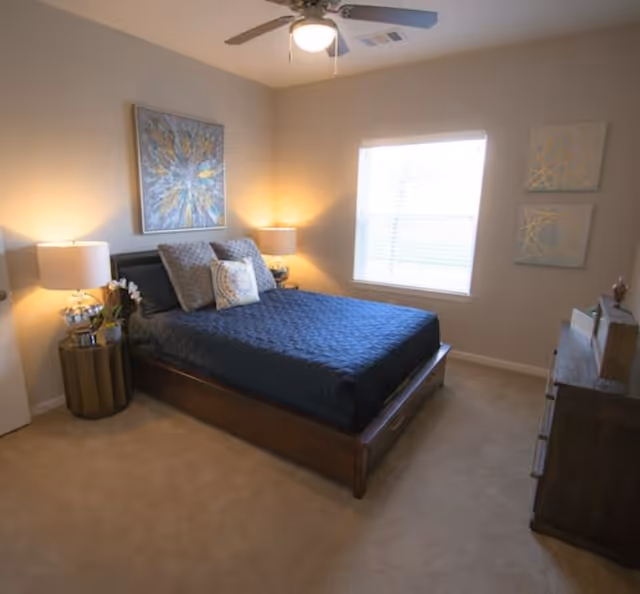A neatly made bed with a dark blue quilt and multiple pillows in a bedroom. The room has beige carpet, a ceiling fan with light, two bedside tables with lamps, a window with blinds, and artwork on the walls. There is also a wooden dresser with decorative items on the right side of the room.