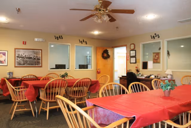 Communal dining/activity room with wooden chairs and tables covered in red tablecloths, a desk area against the wall, and a ceiling fan overhead.