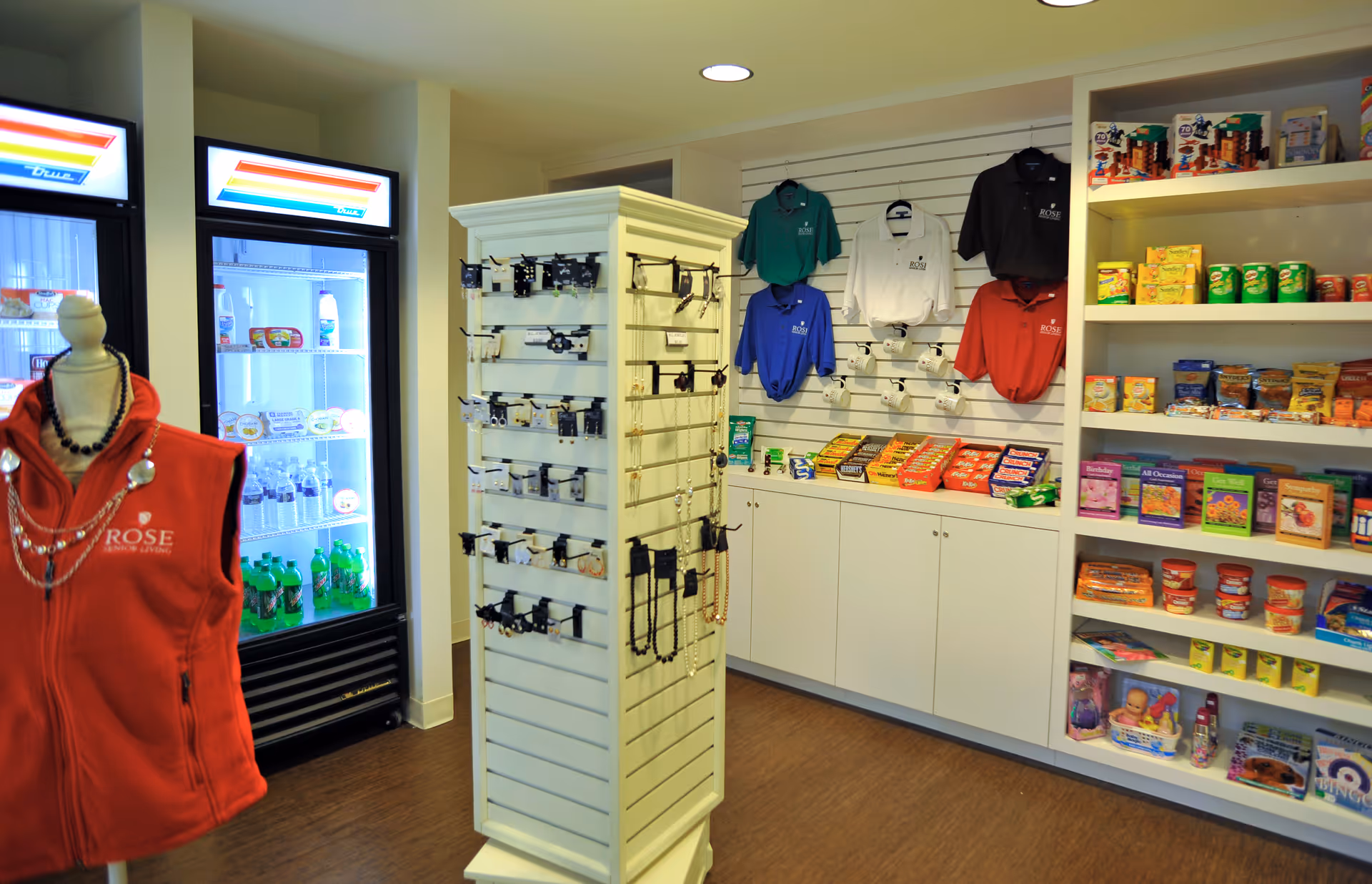 Interior view of a small retail shop area inside Rose Senior Living Clinton Township featuring a mannequin wearing a red vest with the facility's logo, refrigerated coolers stocked with beverages, a display rack with jewelry and accessories, shelves with branded polo shirts, and various snacks and items on white shelves.