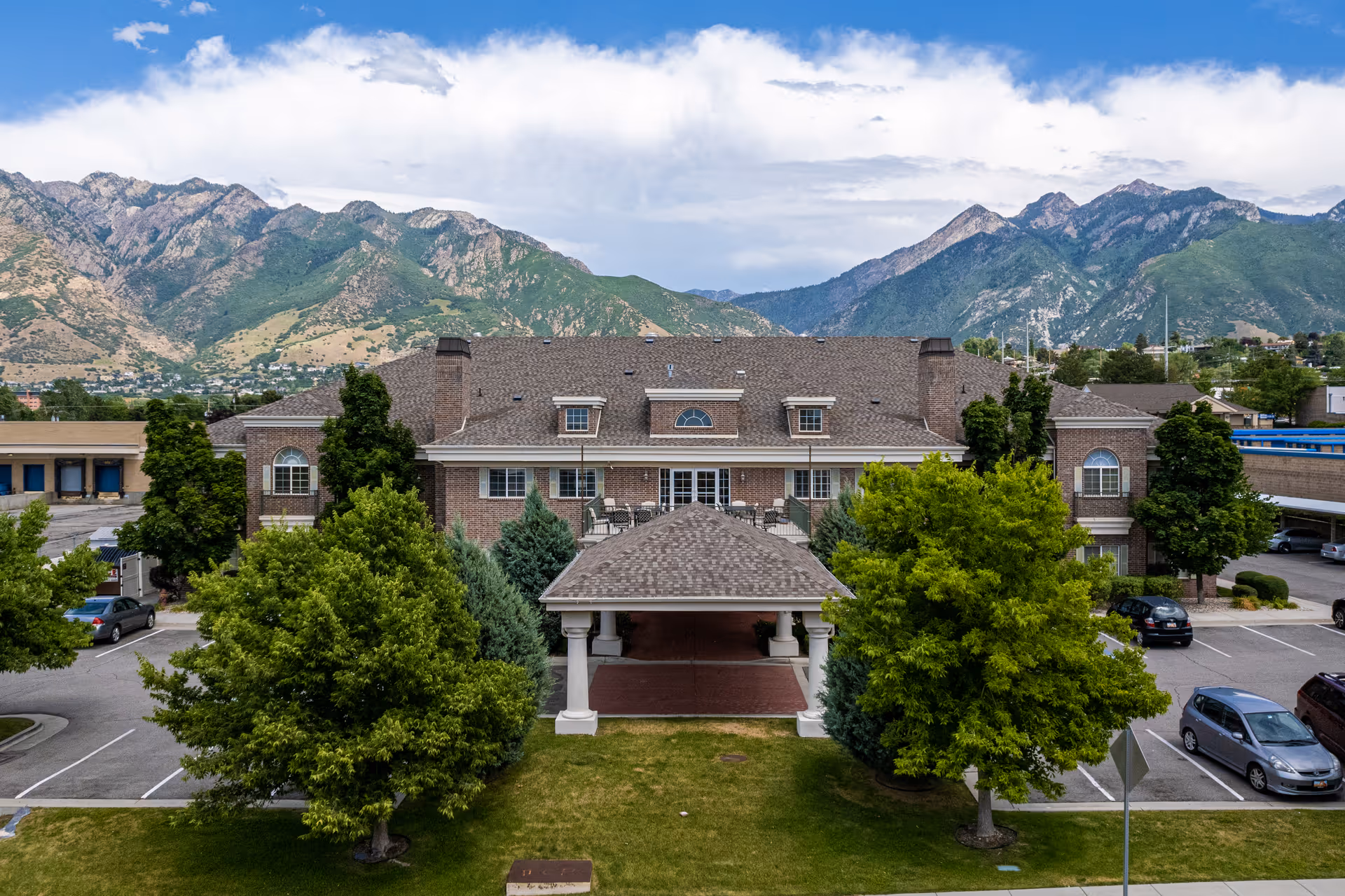 Front exterior view of a large brick building with a covered entrance, surrounded by green trees and parking spaces, with mountains and a partly cloudy sky in the background.