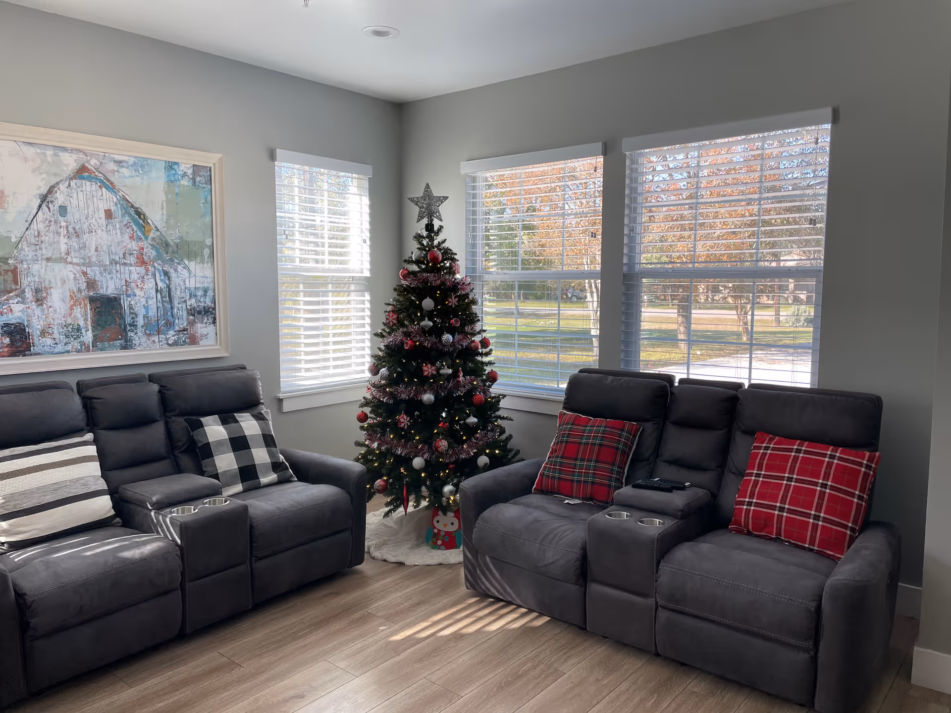 Sunlit living room with two gray reclining sofas, a decorated Christmas tree between windows, and plaid pillows.