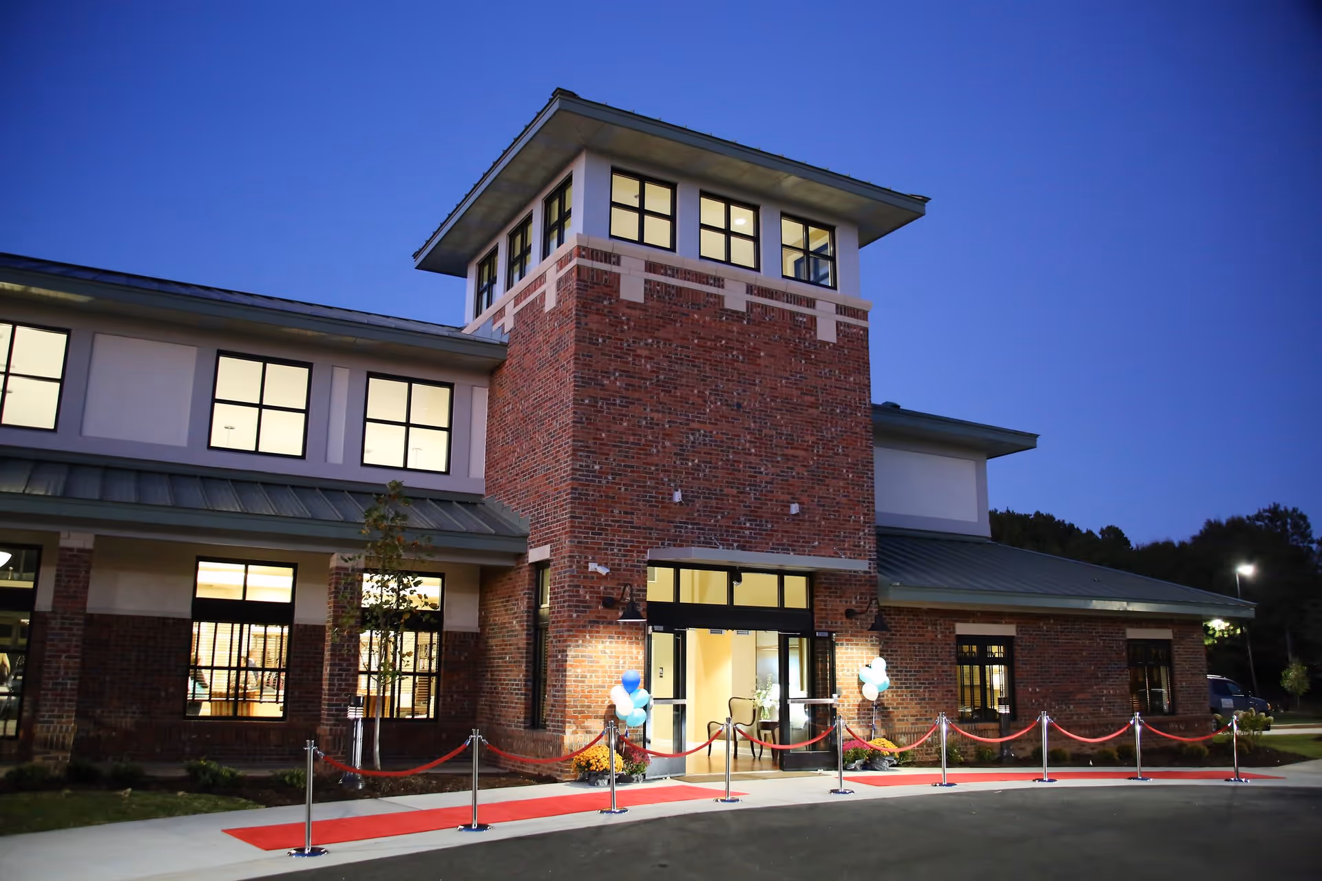 Exterior view of a brick building at dusk with large windows lit from inside. The entrance has glass doors, a red carpet, and stanchions with red ropes. Blue and white balloons are tied near the entrance.