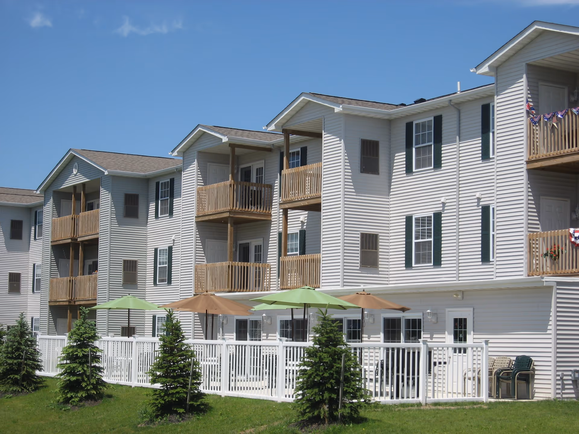 Exterior view of a multi-story senior apartment building with white siding and wooden balconies. There is a fenced patio area with umbrellas and small evergreen trees in front of the building under a clear blue sky.