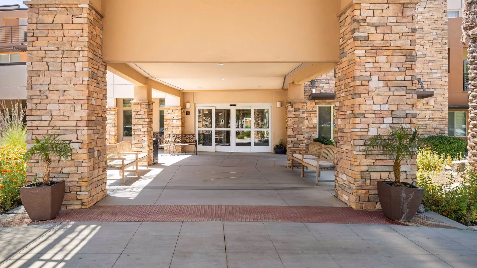 Covered entrance area of a senior living facility with stone pillars, benches on both sides, potted plants, and glass double doors leading inside.