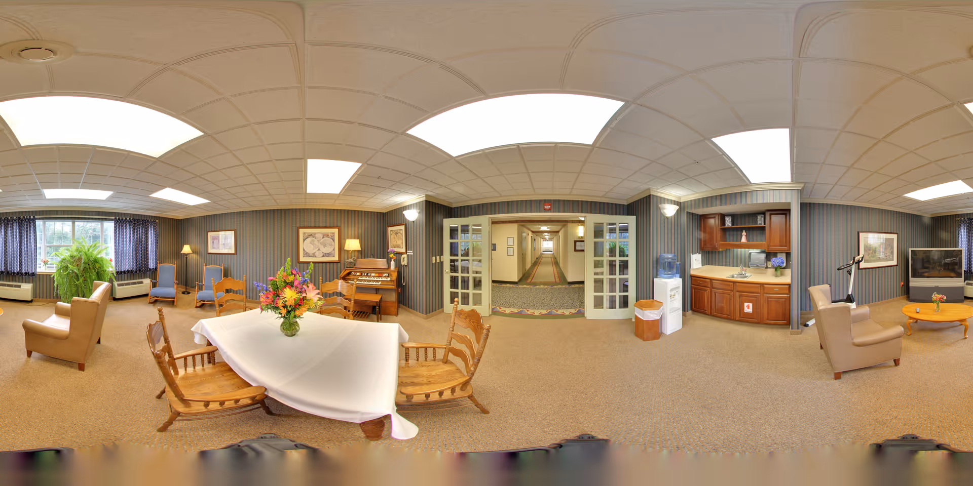 Communal lounge room with seating, a table with a floral centerpiece, piano, kitchenette and TV in a senior living facility.