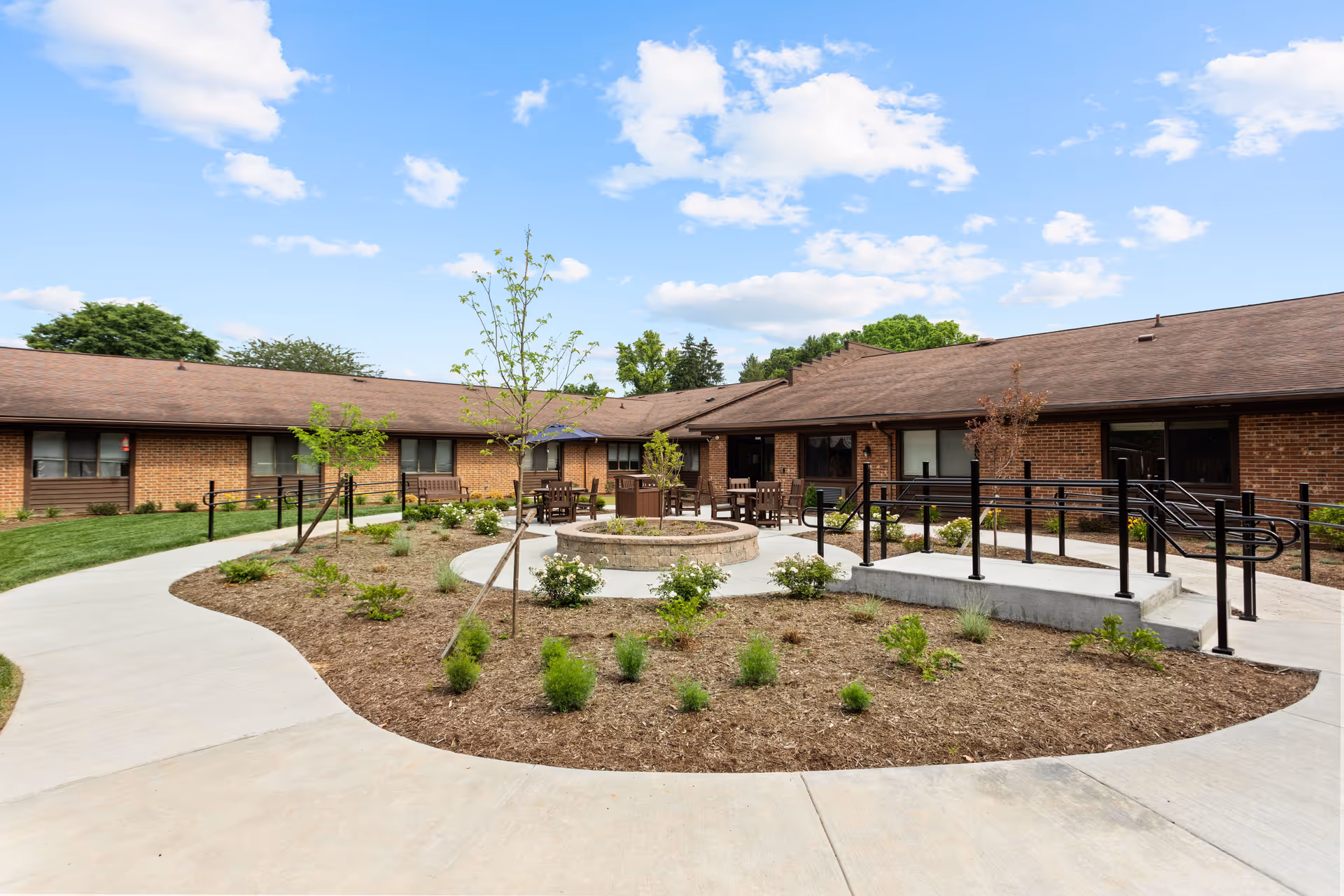 Outdoor courtyard area of a nursing center with a circular stone fire pit surrounded by wooden chairs and tables. The courtyard is landscaped with small plants and young trees, and there are paved walkways and a ramp with black railings leading to the building entrance. The building is a single-story brick structure with a brown roof under a partly cloudy blue sky.