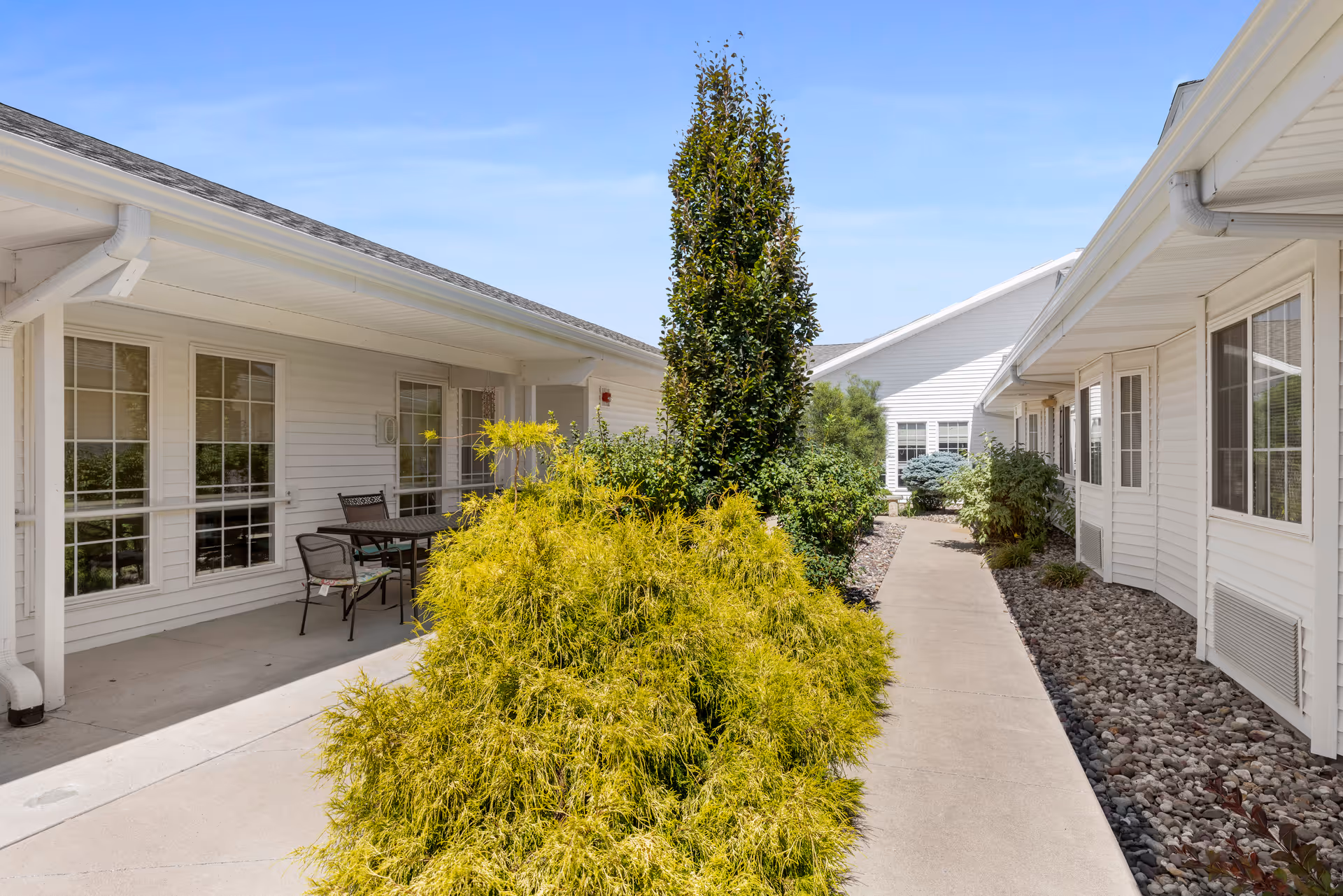 Outdoor walkway between two white buildings with large windows, surrounded by green bushes and plants under a clear blue sky. There is a small patio area with a table and chairs on the left side.