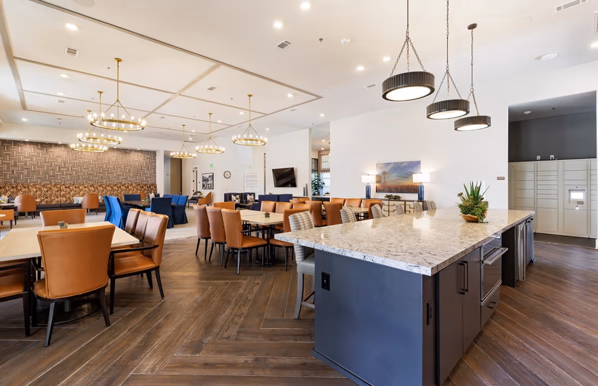 A spacious and modern common area in a senior living facility featuring a large kitchen island with granite countertop and bar stools, multiple dining tables with brown leather chairs, elegant chandeliers hanging from the ceiling, a seating area with blue and brown chairs, and decorative wall art and lamps.