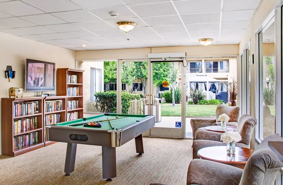 A bright and inviting common room with a pool table in the center, bookshelves filled with books, a wall-mounted television, and comfortable armchairs with small tables holding flower vases. Large glass doors and windows provide a view of a green outdoor garden area.