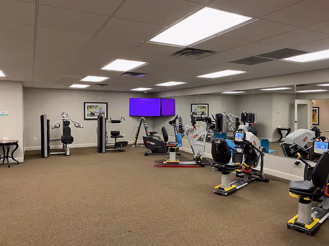 A fitness room in a senior living facility with exercise equipment including stationary bikes, weight machines, and dumbbells. The room has beige carpet, white walls, ceiling lights, and a large mirror on one wall. Two purple screens are mounted on the far wall above a rack of dumbbells.
