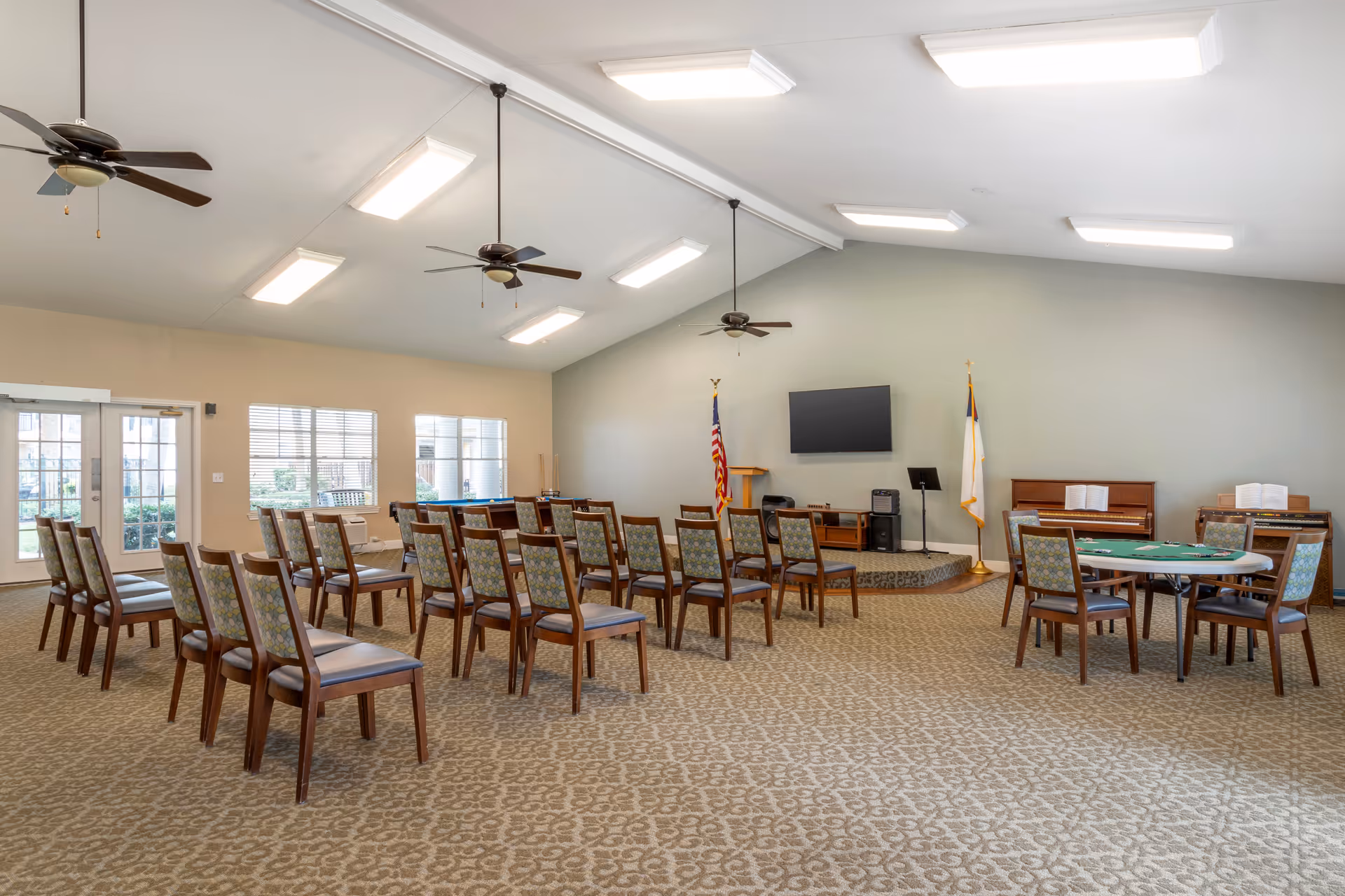 A spacious community room with rows of wooden chairs arranged facing a small stage area. The stage has an American flag, a Christian flag, a podium, a flat-screen TV mounted on the wall, and musical instruments including a piano and an organ. There is a round table with chairs on the right side, and ceiling fans and fluorescent lights are mounted on the high ceiling. Large windows and glass doors allow natural light into the room.