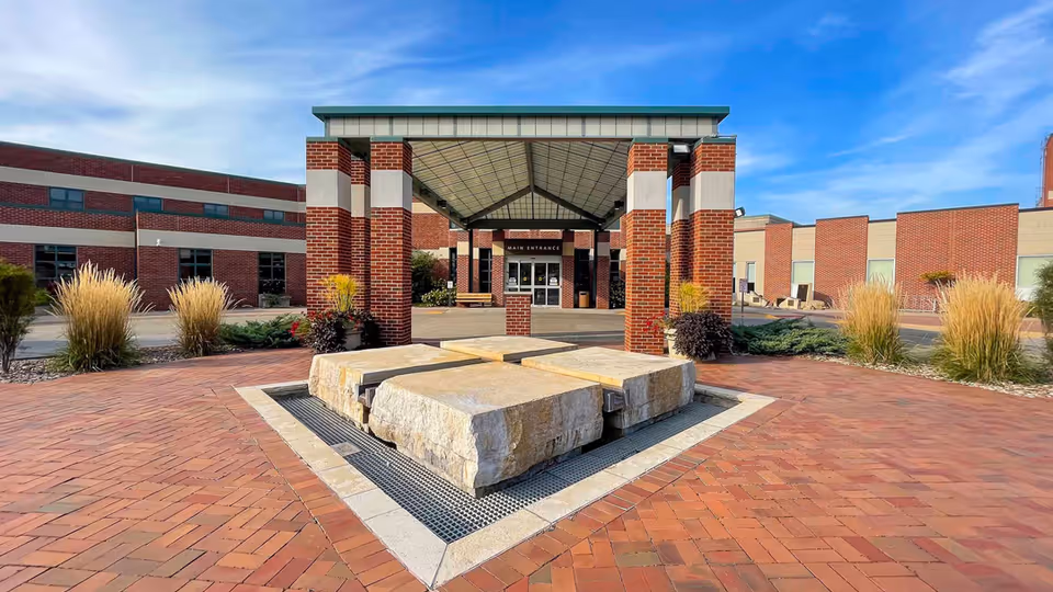 Front entrance of a brick long-term care facility with a covered porte-cochère and a stone water feature in a paved courtyard.