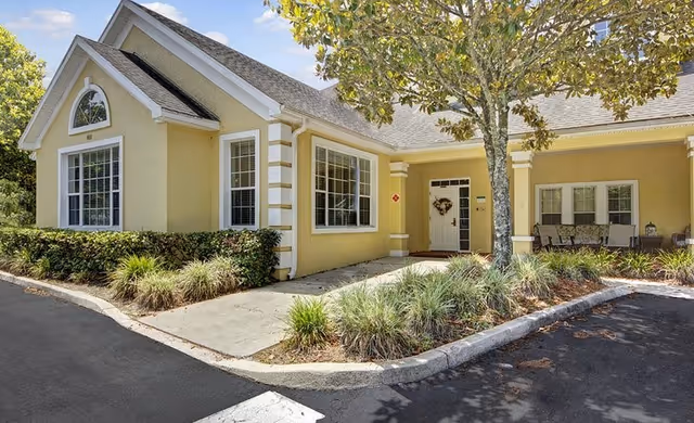 Exterior view of a single-story yellow building with white trim, featuring large windows and a covered entrance. There is a tree and landscaped bushes in front, with a paved driveway and parking area surrounding the building.