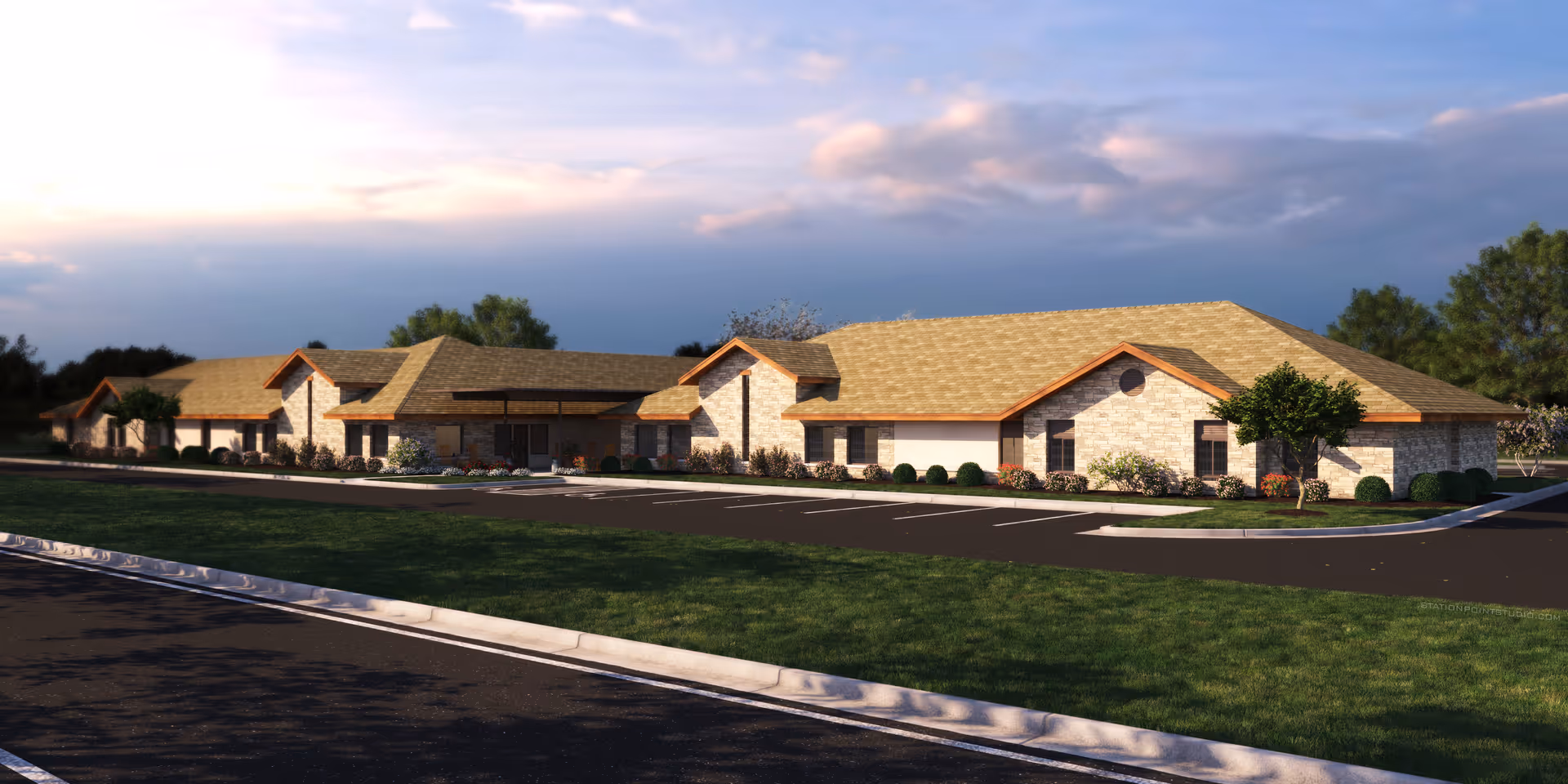 Exterior view of a single-story assisted living facility building with a stone facade and tan shingled roof, surrounded by landscaped greenery and a parking lot under a partly cloudy sky.