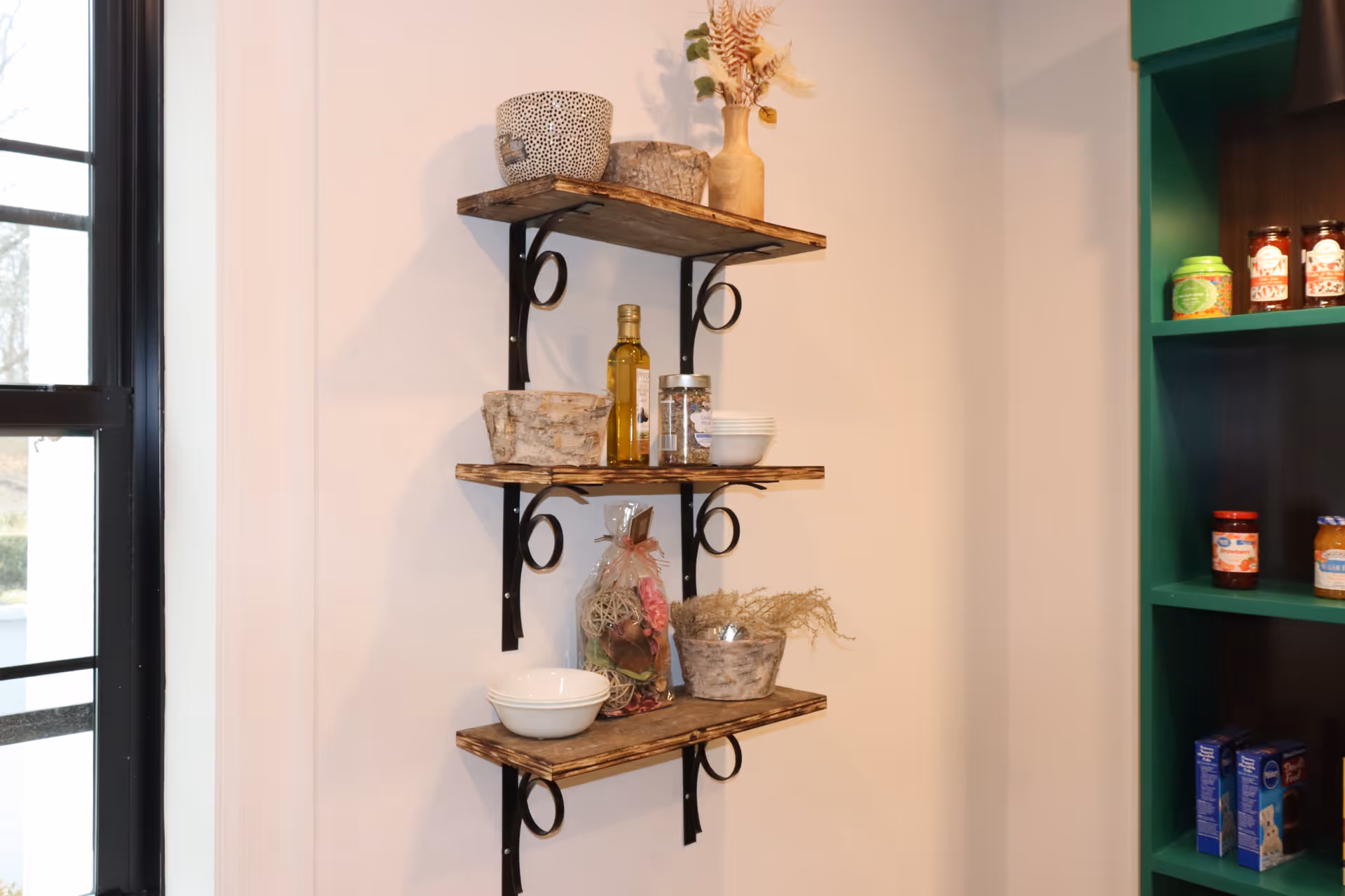 Three wooden wall shelves with black metal brackets mounted on a white wall. The shelves hold various items including decorative baskets, a bottle of olive oil, small bowls, a jar, and a dried flower arrangement. To the right, a green shelving unit contains jars and boxes of food products. A window with black framing is visible on the left side.
