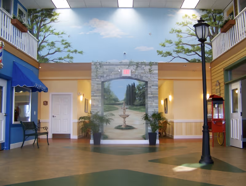 Interior lobby of a senior living facility featuring a painted archway mural with a fountain, a streetlamp, benches and storefront-style facades.