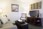 Cozy living room with a sofa, armchair, ottoman, and a TV on a wooden stand beneath wall art.