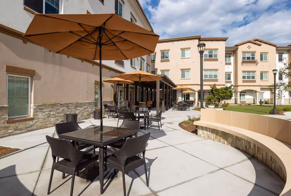 Outdoor patio area at Westmont Of La Mesa featuring multiple tables with umbrellas and chairs on a concrete surface, surrounded by a multi-story building with windows and a small grassy area with plants and lamp posts under a partly cloudy sky.