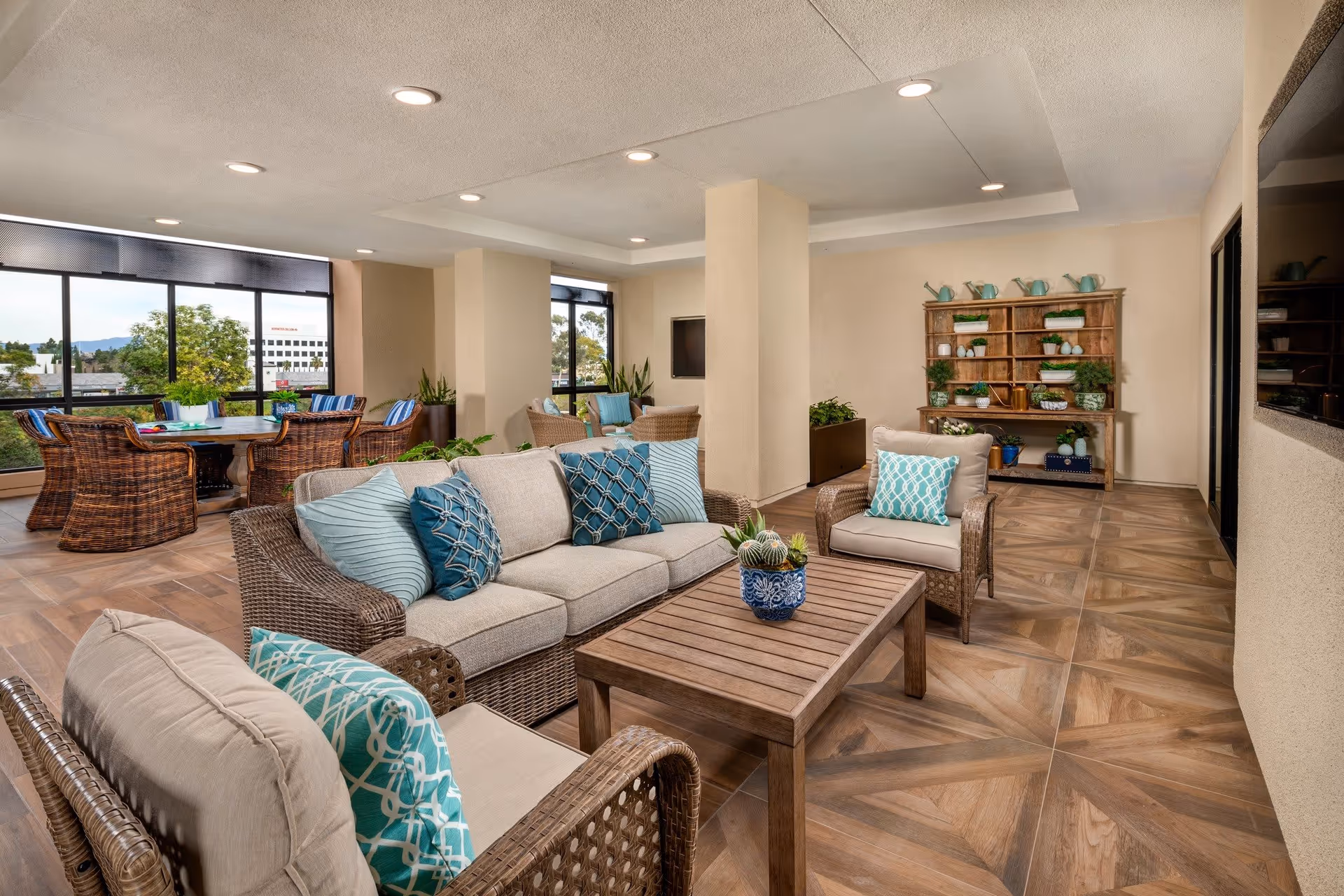 A spacious and well-lit living room area with wicker furniture including a sofa and two armchairs adorned with blue and teal patterned cushions. A wooden coffee table with a small potted plant sits in the center. In the background, there is a wooden shelving unit decorated with various plants and watering cans. Large windows allow natural light to fill the room, and a dining table with wicker chairs is visible near the windows.
