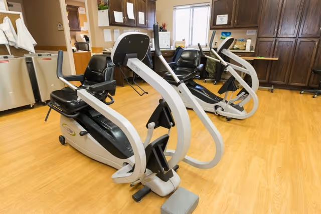 Indoor exercise room with two recumbent stationary bikes on a wooden floor. The room has wooden cabinets and a window letting in natural light.