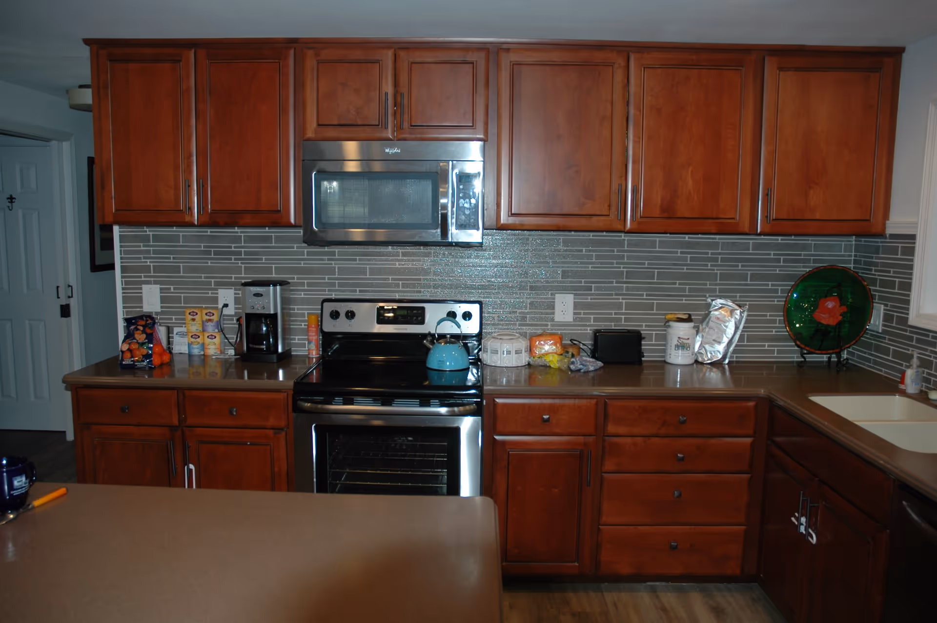 A kitchen with wooden cabinets and drawers, a stainless steel microwave and oven, a coffee maker, a blue kettle on the stove, various food items on the countertop, and a green decorative plate near the sink.