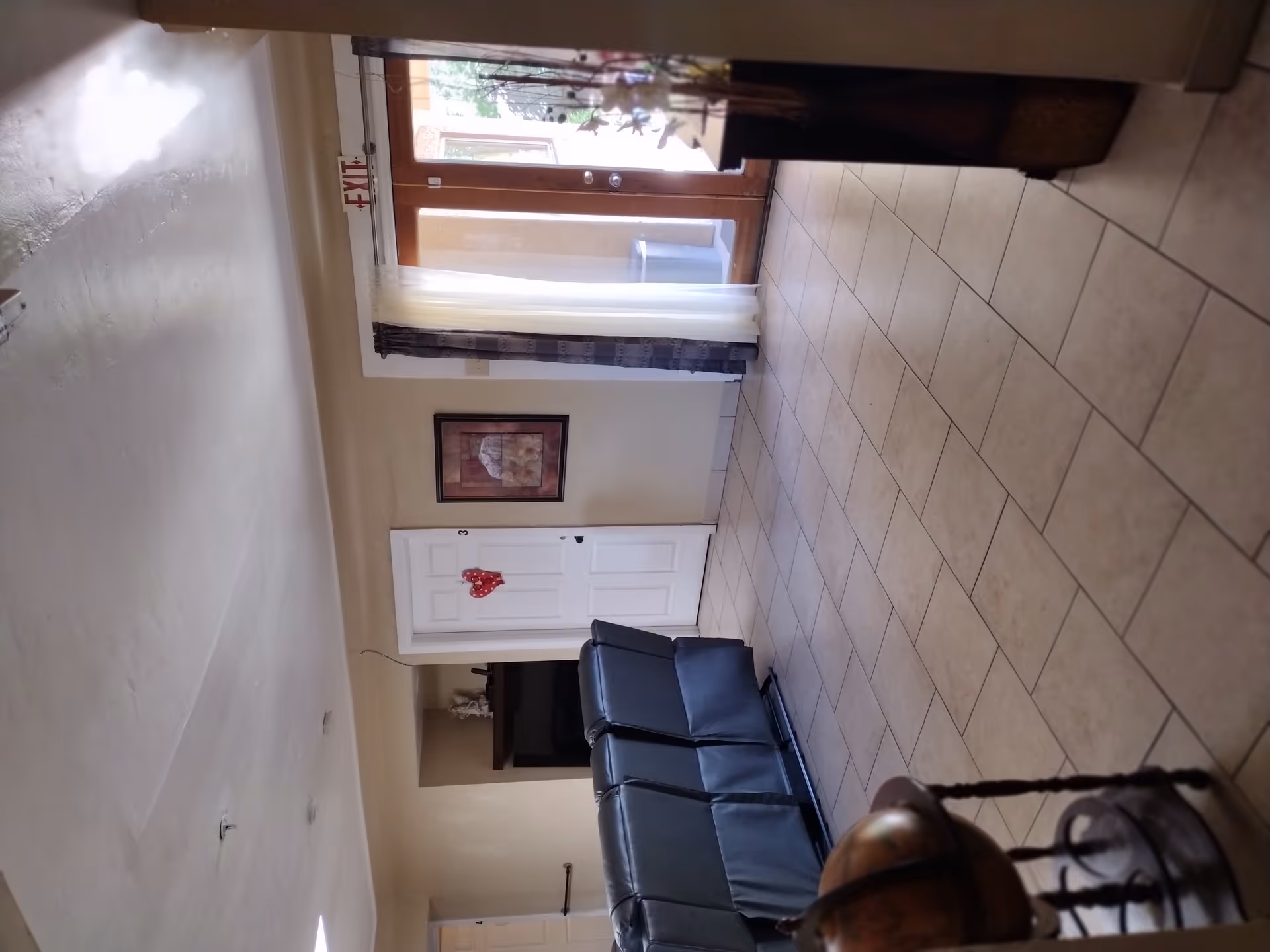Interior view of a living room area in an assisted living home with beige tiled floor, cream-colored walls, a row of black recliner chairs facing away, a white door with a red heart decoration, a framed picture on the wall, and glass double doors with curtains leading outside. A globe on a stand is partially visible in the foreground.