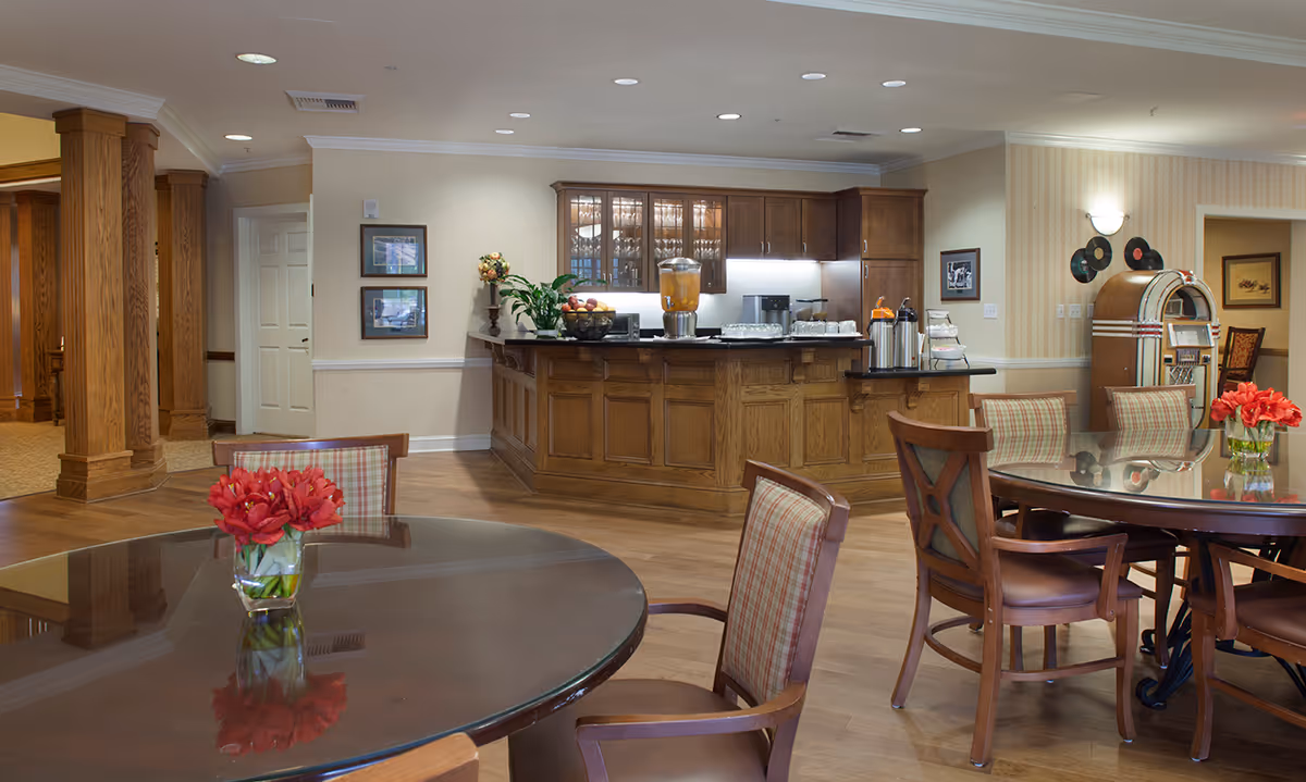 Communal dining area with round glass-topped tables, upholstered chairs, a wooden service counter and a jukebox in the background.