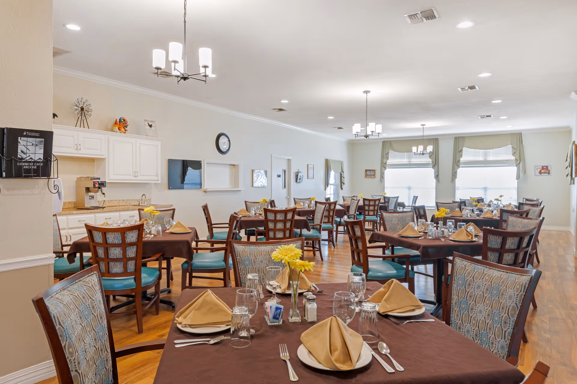 A spacious dining room in a senior living facility with multiple tables covered in brown tablecloths, each set with beige napkins, plates, glasses, and silverware. The chairs have patterned upholstery and teal seats. The room has wooden flooring, large windows with valances, and ceiling lights. A small counter area with cabinets and a coffee machine is visible on the left side.