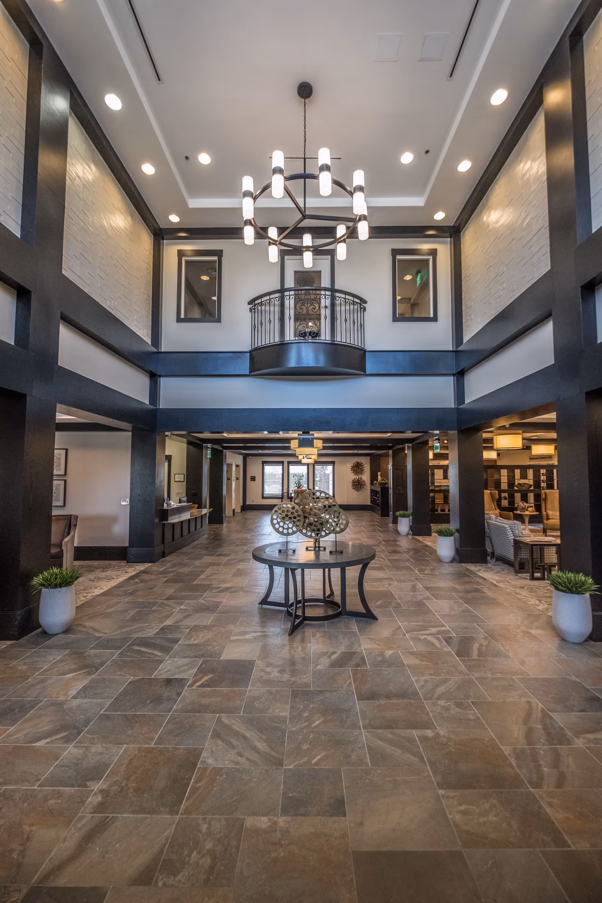 Spacious and modern lobby area with high ceilings, a large chandelier, tiled floor, and a round table with decorative items in the center. The space features dark wood beams, light-colored walls, potted plants, and seating areas on the sides.