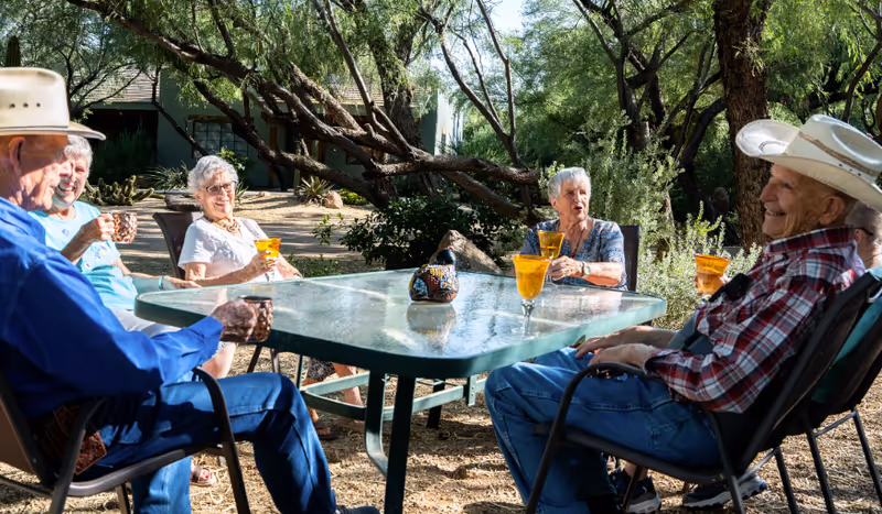A group of seniors sitting around an outdoor table under trees, enjoying drinks and conversation.