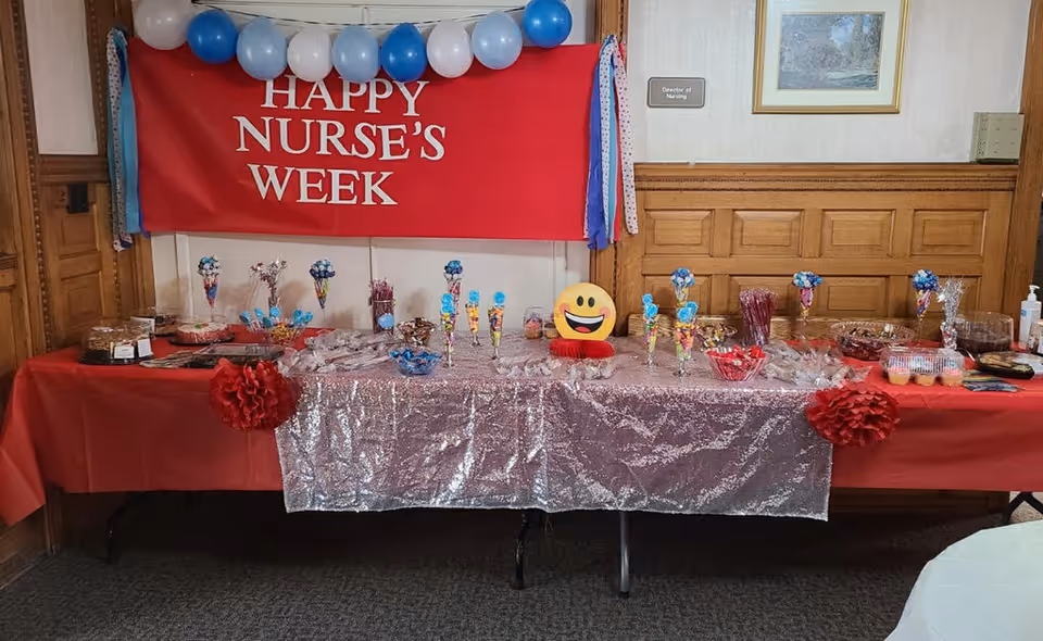 A decorated table set up for a celebration with a red banner reading 'HAPPY NURSE'S WEEK' hanging on the wall behind it. The table is covered with a red and silver sequin tablecloth and displays various snacks and treats, including cupcakes, candy, and other desserts. Blue, white, and light blue balloons are arranged above the banner. The setting appears to be inside a room with wood-paneled walls and a sign indicating 'Director of Nursing'.