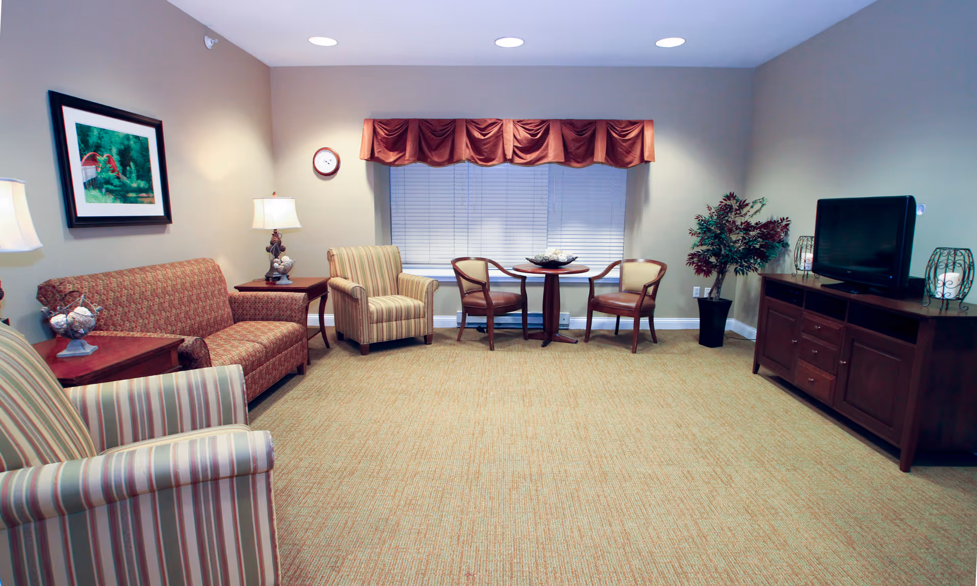 Well-lit living room/lounge with sofas and armchairs arranged around a small table, a TV on a wooden cabinet, and a window with a valance.