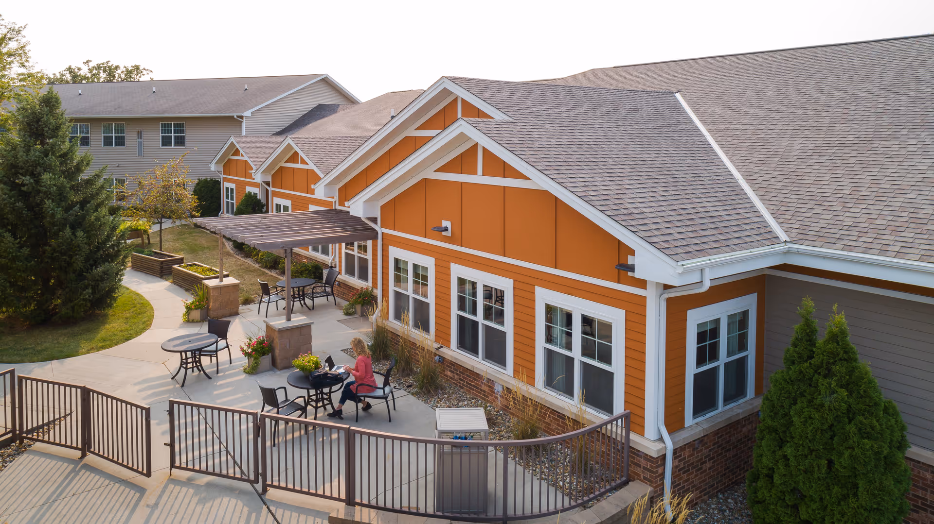 Outdoor patio area at Newton Village with tables and chairs, a woman sitting at one table, surrounded by greenery and a pathway leading to the building entrance.