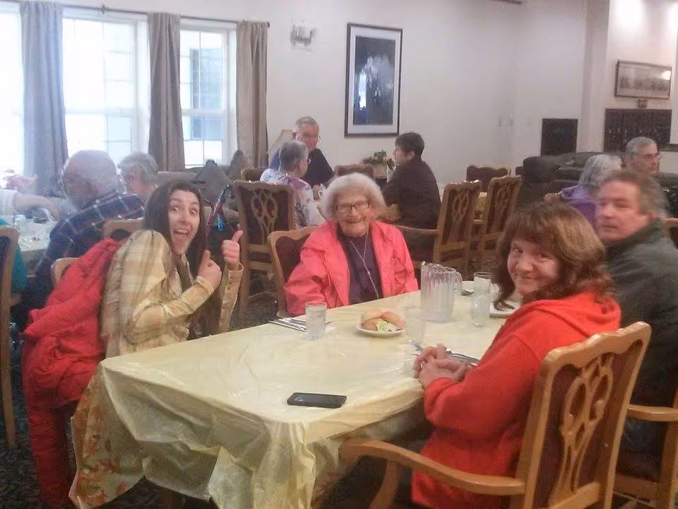 A group of elderly and middle-aged people sitting around tables in a dining room, smiling and engaging with each other. The room has large windows with curtains, wooden chairs, and tables covered with plastic tablecloths. There are pitchers of water and glasses on the tables.
