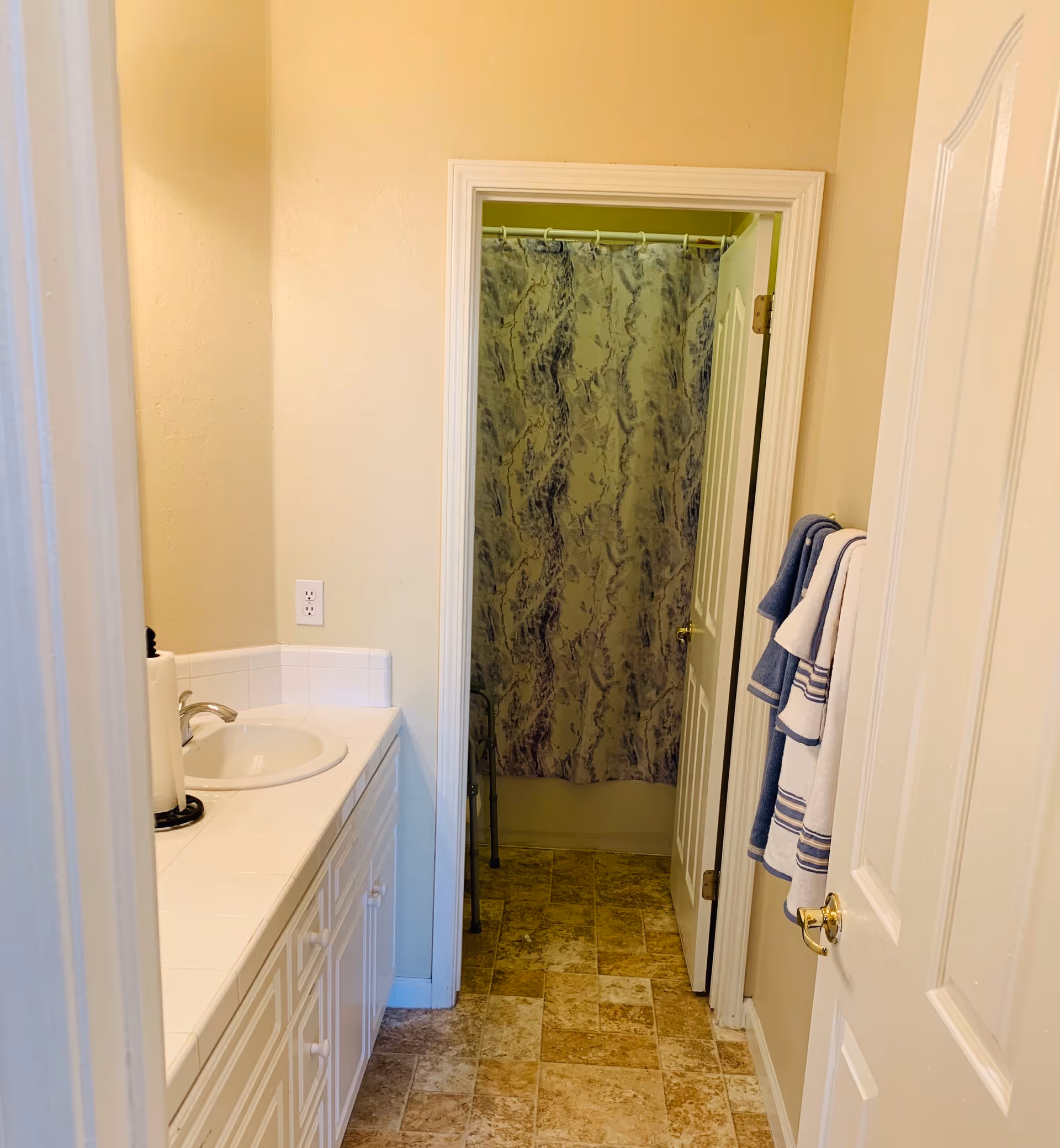 A bathroom with a white sink and countertop on the left side, a paper towel holder next to the sink, beige walls, and a tiled floor. A doorway leads to a shower area with a marbled shower curtain and a walker visible inside. On the right wall, there are blue and white striped towels hanging on a towel rack.