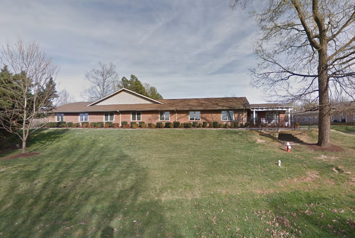 Front exterior of The Oaks Of Alamance, a single-story brick senior living facility with manicured lawn.