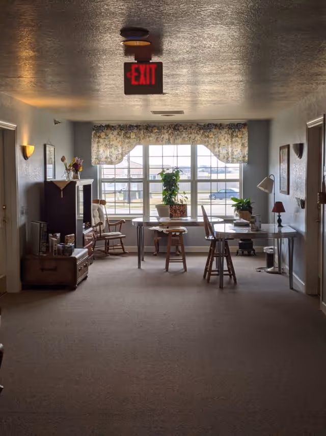A cozy interior room with a large window covered by floral curtains letting in natural light. In front of the window, there is a table with two wooden chairs and potted plants on top. To the left, there is a rocking chair and a wooden cabinet with decorative items and flowers. The walls are light-colored with framed pictures and wall lamps. A red exit sign is mounted on the ceiling.