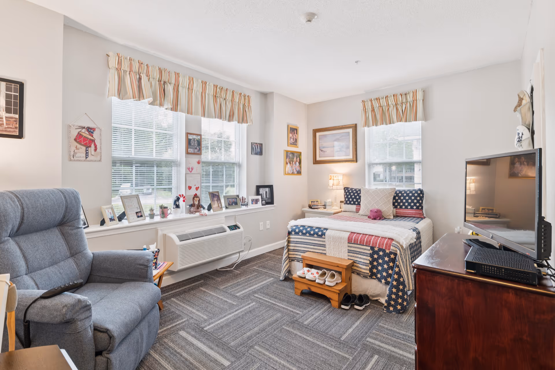 Well-lit bedroom with a patriotic quilted bed, a recliner, TV on a dresser, and a window ledge displaying photos and decorations.