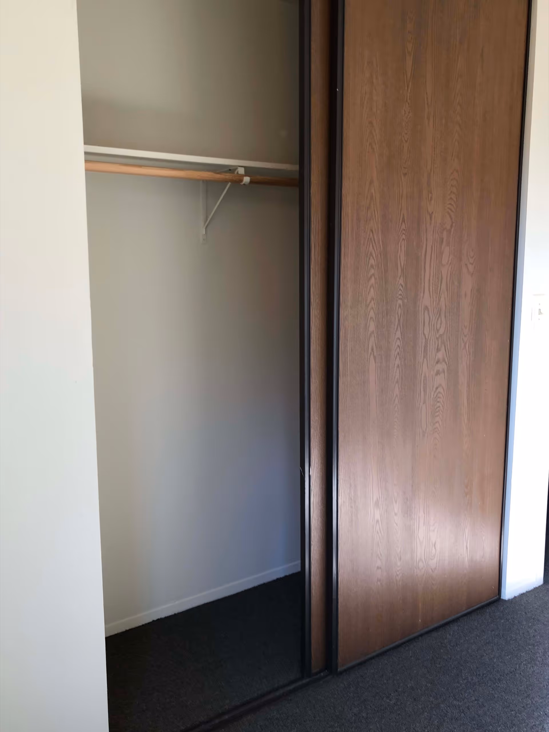 An empty closet with a wooden sliding door partially open, showing a wooden hanging rod inside. The closet walls are white and the floor is carpeted in dark gray.