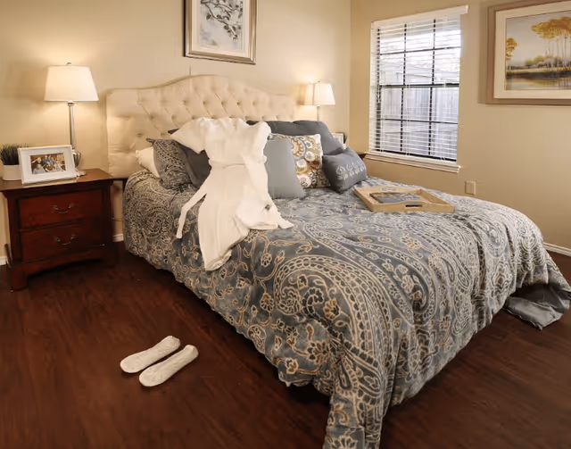 A cozy bedroom with a large bed covered in a patterned blue and beige comforter, multiple pillows, and a white robe draped over it. There is a wooden nightstand with a lamp and a framed photo on the left side of the bed. A window with blinds is on the right wall, and a framed painting hangs beside it. A pair of white slippers is placed on the wooden floor near the bed.