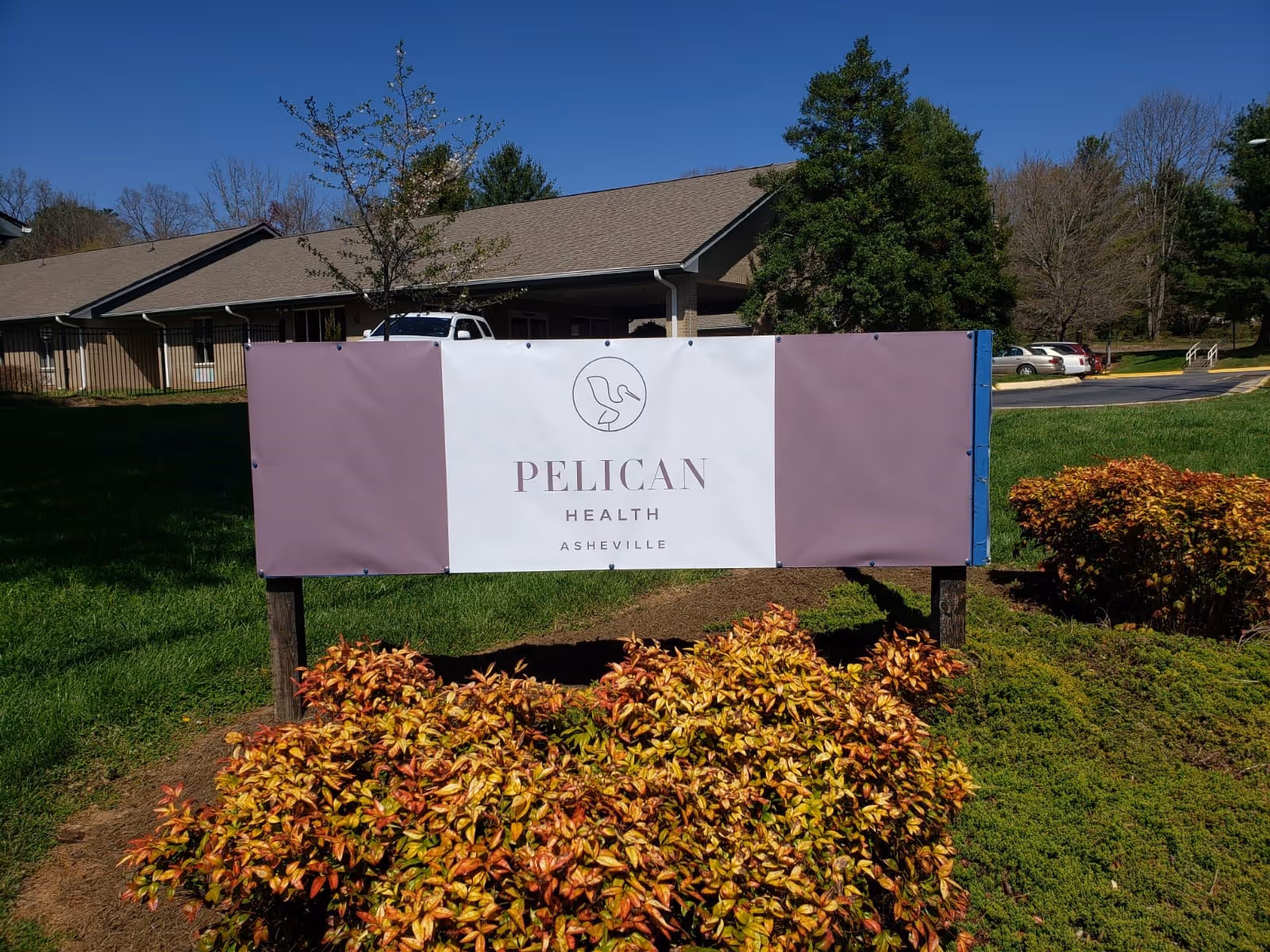 Outdoor view of a sign for Pelican Health Asheville in front of a building with a sloped roof, surrounded by green grass, bushes with reddish leaves, and trees under a clear blue sky.