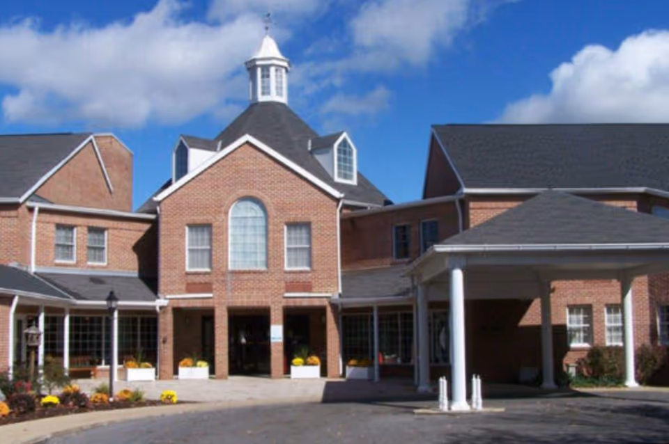 Front exterior view of a large brick building with a peaked roof and a cupola on top, featuring multiple windows and a covered entrance supported by white columns under a blue sky with some clouds.
