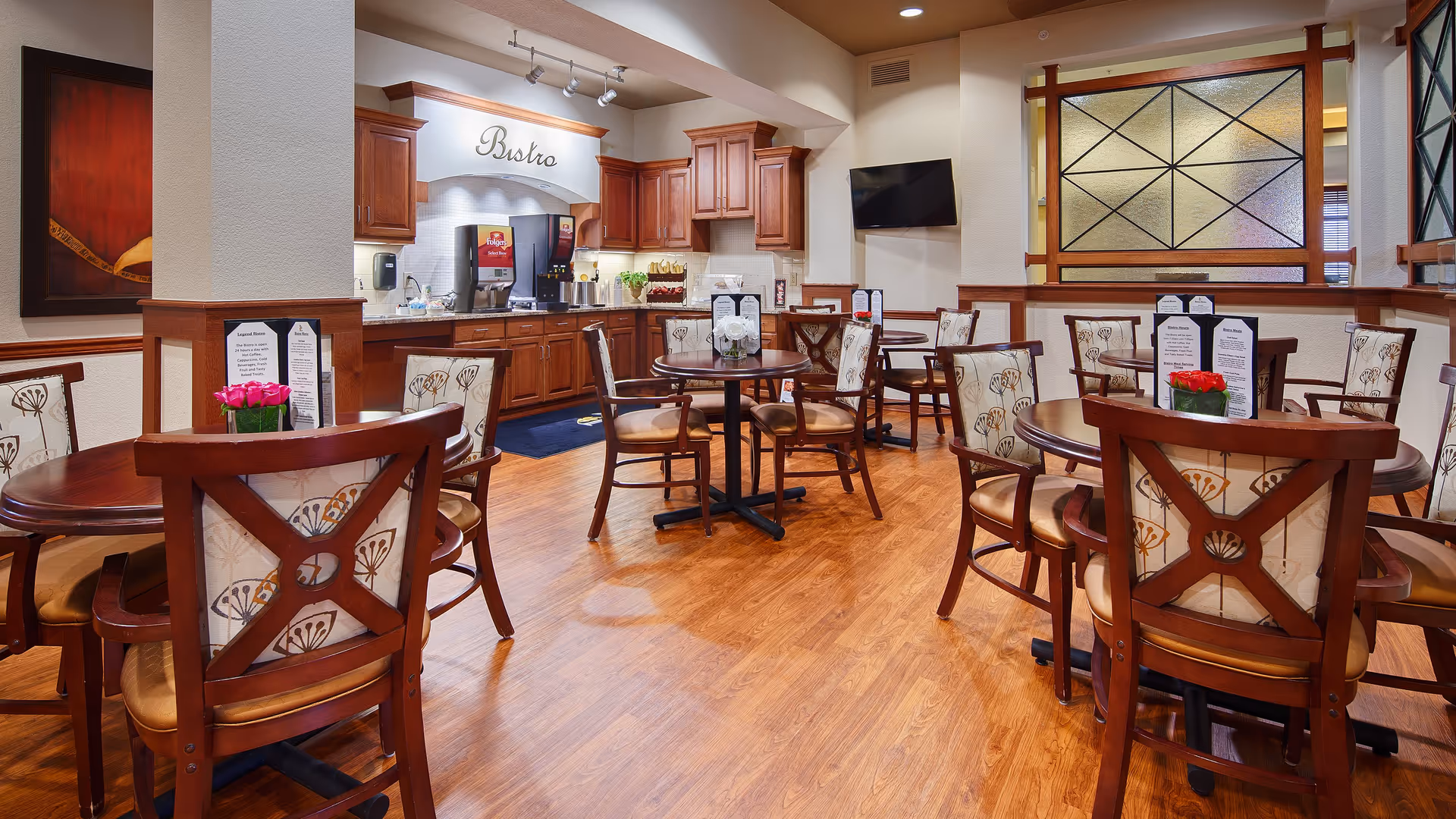 Interior view of a dining area in a senior living facility with wooden floors, round wooden tables, and cushioned chairs with patterned backs. The back wall features a beverage station labeled 'Bistro' with wooden cabinets and a soda dispenser. Menus and small flower arrangements are placed on the tables. A flat-screen TV is mounted on the wall to the right.
