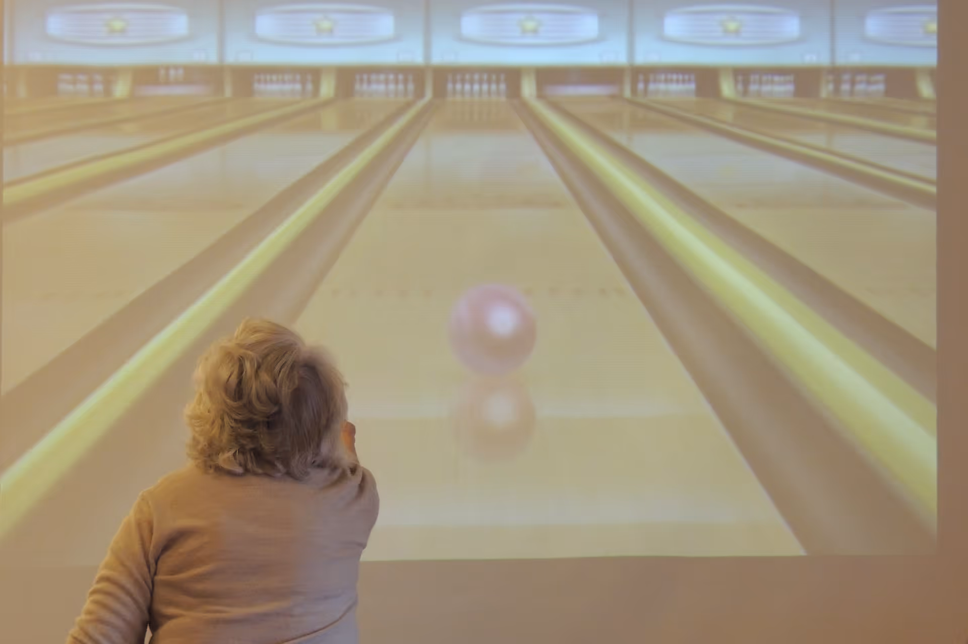 An elderly person with gray hair seen from behind is playing a virtual bowling game projected on a wall, showing bowling lanes and a bowling ball rolling down the lane.