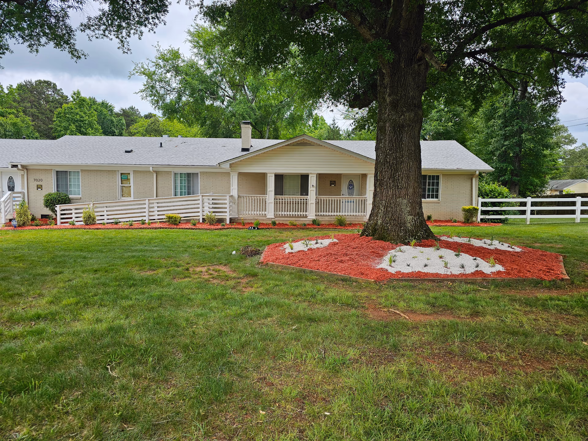 Single-story building with a light-colored exterior and a gray roof, featuring a front porch with railings and a ramp. In front of the building is a large tree surrounded by a landscaped area with red mulch and white stones. The lawn is green and well-maintained, with trees and greenery in the background under a cloudy sky.