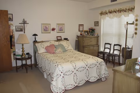 A cozy bedroom with a double bed covered in a white quilt with a floral pattern and several pillows. There is a wooden nightstand with a lamp and some items on it to the left of the bed. On the wall above the bed are three framed pictures. To the right of the bed is a wooden dresser with decorative items and flowers on top. Two wooden chairs are placed near a window with blinds and a floral valance. The room has beige carpet and light-colored walls.