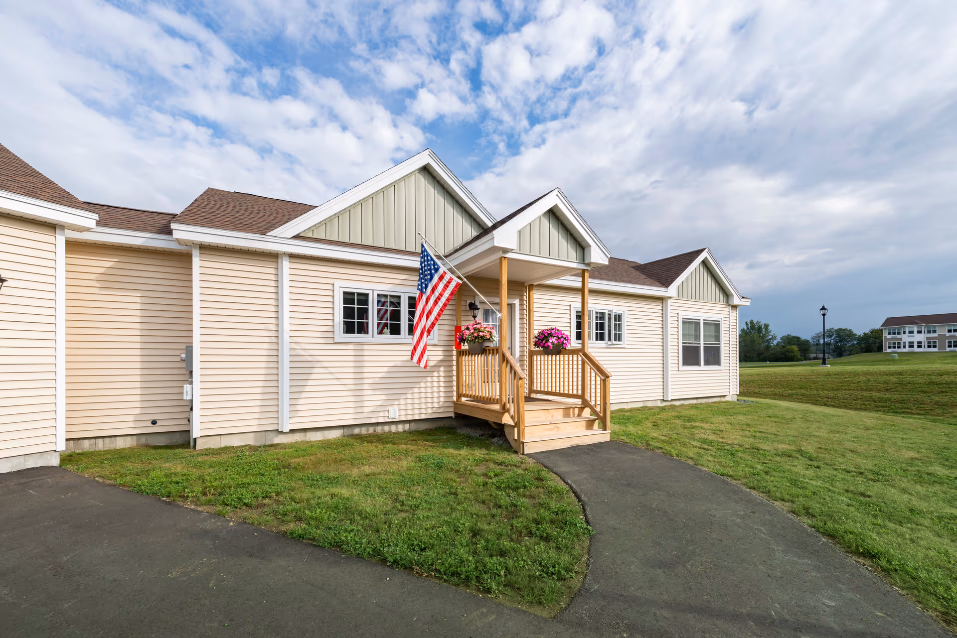 Exterior view of a single-story building with beige siding and a small covered porch with wooden railings. An American flag and flower pots with pink flowers are displayed on the porch. The building is surrounded by green grass and a paved walkway leads to the porch. The sky is partly cloudy.